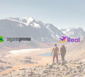 Casal de mochileiros aprecia a vista das montanhas nevadas durante uma trilha. Em primeiro plano, logo Seguros Promo e Real Seguro Viagem