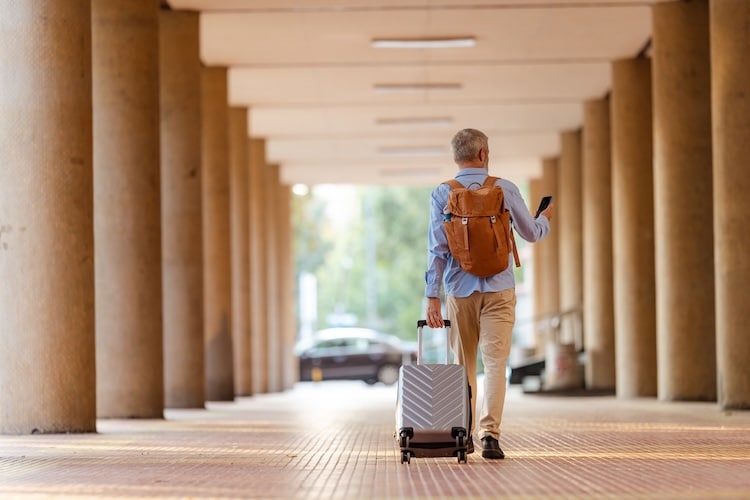 Homem caminhando em um corredor coberto de aeroporto, puxando uma mala de mão e olhando o celular.