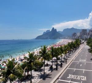 Vista da praia de Ipanema no Rio de Janeiro com banhistas, coqueiros e o Morro Dois Irmãos ao fundo.